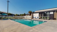 Located at 100 Swift Blvd in Goose Creek, South Carolina, this single-level outdoor amenity area centers around a well-maintained rectangular swimming pool with clear, blue-green water. The pool is framed by a spacious concrete sun deck that provides ample room for lounging, with multiple sunbathing chairs positioned to take full advantage of the South Carolina sunshine. A dedicated seating area features a round table shaded by a teal umbrella, accompanied by comfortable chairs — ideal for relaxing poolside. The entire area is enclosed by a clean white vinyl privacy fence, offering a sense of seclusion while maintaining an open, airy feel under the expansive sky. Lush green lawns and mature palm trees add a tropical ambiance, enhancing the resort-like atmosphere of the space. A compact pool house structure is situated adjacent to the pool, providing functional support facilities. The surrounding landscape includes well-kept grassy areas visible through the fence, contributing to the verdant, welcoming character of the property. Overall, this outdoor space is thoughtfully designed for leisure and recreation, offering a comfortable and attractive environment suited to the warm climate of coastal South Carolina.