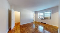 This single-floor unit is characterized by warm parquet hardwood flooring that runs throughout the main living areas and bedroom, lending a classic, timeless aesthetic to the space. The living area is generously proportioned, featuring neutral white walls, white-trimmed baseboards, and windows fitted with horizontal blinds that allow natural light to fill the room. A wall-mounted electrical panel and window air conditioning unit are notable functional elements. The space connects openly to a separate bedroom, which features the same parquet flooring, a closet, and clean, understated finishes.

The kitchen is outfitted with warm oak cabinetry extending both above and below the countertops, providing ample storage. A stainless steel gas range and a double-basin sink complement the light-toned countertops, while light tile flooring differentiates this area from the rest of the unit. An adjacent transitional space with matching tile flooring and a window offers additional utility.

The bathroom maintains a classic design with white wall tiles accented by a dark contrasting border, a built-in bathtub with overhead shower, a white vanity with sink, and a mirrored medicine cabinet, creating a clean and functional environment throughout.