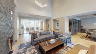 This contemporary amenity space is thoughtfully designed to deliver a refined and welcoming environment across a single open floor. The lounge area commands immediate attention with its soaring double-height ceilings, an eye-catching sculptural LED chandelier, and floor-to-ceiling windows that flood the interior with natural light and frame views of lush outdoor greenery. A dramatic stacked-stone accent wall serves as the focal point, housing a sleek linear fireplace and a wall-mounted television, while custom built-in shelving with warm wood accents adds both function and visual depth. Stylish seating arrangements, anchored by a patterned area rug and a solid wood coffee table, create an inviting atmosphere ideal for gathering or relaxing. Polished concrete flooring flows seamlessly throughout, lending an industrial-modern character to the space. The kitchenette area features clean-lined cabinetry, light stone countertops, a built-in microwave, and large-format wall tiles, providing a practical yet elegant service zone. Glass-paneled doors connect to adjacent areas, ensuring an open, airy feel. Curated artwork and carefully placed greenery complete the aesthetic, making this a polished and functional communal space.