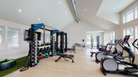 Located at 4550 Atrium Ct in Wilmington, NC, this single-floor fitness facility is thoughtfully designed to accommodate a full range of training disciplines. The space is defined by soaring vaulted ceilings with recessed lighting and exposed ductwork that lends an industrial-chic aesthetic, all set against crisp white walls and light hardwood-style flooring that creates a bright, energizing atmosphere.

The strength training zone is anchored by a robust power rack system outfitted with a comprehensive dumbbell rack, kettlebells, medicine balls, and adjustable benches, providing everything needed for a complete resistance workout. Adjacent to this area, a dedicated turf section with artificial grass supports functional training activities, featuring a tire flip station, agility cones, plyo boxes, and a wall-mounted flat-screen TV for guided programming.

The cardio zone is generously stocked with multiple treadmills, elliptical machines, and stationary bikes — including a spin bike — all positioned near large windows that frame views of the outdoor pool area, filling the space with natural light and visual connection to the outdoors. A cable machine and additional free weight stations round out the strength offerings. Practical amenities include a wall-mounted water fountain station, ensuring convenience during intense sessions. The facility's open floor plan and high ceilings create a spacious, motivating environment suited to individual and group training alike.