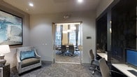 This single-floor professional space combines functionality and refined design to create an ideal environment for meetings and focused work. The centerpiece is a glass-walled conference room, accessible through a sleek framed glass door that allows natural light to flow between spaces while maintaining a sense of enclosure. Inside the conference room, a large conference table is surrounded by ergonomic high-back chairs, accommodating a sizable group comfortably. Elegant pendant light fixtures hang overhead, providing warm, focused illumination, while floor-length draping curtains frame the windows, which offer views of the surrounding exterior. The patterned carpet throughout adds texture and visual interest while tying the rooms together cohesively. Adjacent to the conference room, a secondary work area features a built-in desk surface, additional task seating, and a large wall-mounted display screen, suitable for presentations or individual work. A cushioned accent chair with a teal throw pillow adds a touch of comfort near the entrance. The neutral color palette of soft grays and warm tones contributes to a calm, professional ambiance throughout the space.