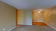 This single-floor space presents a straightforward, open layout divided into two distinct areas distinguished by their flooring. The front section features wall-to-wall carpeting in a neutral tone, providing a comfortable, soft underfoot feel, while the adjacent area transitions to warm hardwood flooring, adding a touch of natural character to the space. The walls throughout are finished in a warm beige tone, contributing to an inviting, neutral atmosphere that serves as a versatile backdrop for various furnishings and décor styles. White baseboards run along the perimeter, offering a clean contrast to the warmer wall tones. A ceiling fan with an integrated light fixture is installed in the hardwood-floored section, providing both air circulation and ambient lighting. The textured white ceiling adds subtle depth overhead. Multiple electrical outlets are distributed along the walls, ensuring practical functionality throughout. The overall design is modest and unfussy, making the space highly adaptable for a range of uses.
