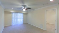 This single-floor residential space offers a clean, functional layout designed for comfortable everyday living. The main living area is a standout feature, boasting wall-to-wall carpeting that adds warmth underfoot and a sense of coziness throughout. Crown molding lines the perimeter of the ceiling, lending the room a refined, polished appearance that elevates the overall aesthetic.

A ceiling fan with an integrated light fixture is centrally mounted, providing both practical air circulation and ambient lighting. The room benefits from generous natural light, courtesy of a large sliding glass door dressed with vertical blinds, which also offers the potential for outdoor access and views of the surrounding exterior. The neutral, light-toned walls create a versatile backdrop suited to a variety of interior styles and furnishings.

A floor-level air vent is positioned along the baseboard, indicating a built-in climate control system for year-round comfort. White baseboards frame the room cleanly, complementing the crisp crown molding above. The overall ambiance is bright, airy, and well-maintained, presenting a move-in-ready space that balances simplicity with thoughtful architectural detailing throughout its single level.
