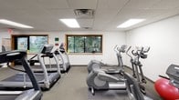 This fitness center is a comprehensively equipped single-floor facility designed to support a wide range of workout routines. The space is organized into distinct functional zones, each tailored to a specific type of exercise. The cardio area features multiple treadmills, elliptical trainers, and a recumbent stationary bike, all arranged to maximize floor space and usability. A wall-mounted flat-screen television is positioned for easy viewing during cardio sessions.

The strength training section includes a multi-function cable machine, adjustable weight benches, and a dumbbell rack with a full set of free weights. Rubber floor mats provide traction and protection in these high-use zones. A large wall mirror expands the visual depth of the space and allows users to monitor their form. Additional accessories such as stability balls, resistance equipment, and a storage rack round out the offerings.

The facility features a drop ceiling with recessed panel lighting that provides even, consistent illumination throughout. Carpeted flooring covers the general circulation areas, while rubber matting is used beneath equipment. Windows along one wall allow natural light to enter and offer views of surrounding greenery. A water fountain and open shelving unit are also present, adding everyday convenience to the space.