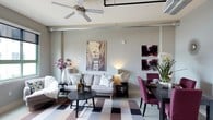 This single-floor urban residence showcases a thoughtfully designed open-concept layout that seamlessly integrates the living and dining areas into one cohesive, functional space. The interior reflects a modern aesthetic with industrial undertones, evident in the exposed ductwork and ceiling-mounted track-style elements that contrast tastefully with the soft, neutral wall tones.

The living area is anchored by a light gray sofa adorned with patterned throw pillows, complemented by a bold mustard yellow accent chair that introduces a lively pop of color. A large geometric-patterned area rug in muted tones of mauve, gray, and cream grounds the seating arrangement around a sleek, round dark coffee table. A ceiling fan overhead provides both functionality and a finishing architectural touch.

Large windows flood the space with natural light and frame impressive city skyline views, enhancing the open, airy atmosphere. The dining area features a dark wood table paired with rich plum-colored upholstered chairs, creating an elegant contrast against the neutral backdrop. Curated wall art, including a European cityscape print and bold floral canvases, adds personality and visual interest throughout. Lush greenery and floral arrangements bring warmth and life to the overall ambiance.