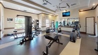 Located at 5225 Zachary Grove in Colorado Springs, CO, this single-floor fitness center is thoughtfully designed to support a comprehensive range of workout routines. The facility is divided into several distinct zones, each tailored to specific training needs.

The cardio area houses multiple treadmills and elliptical machines, a stationary bike, and a rowing machine, all arranged beneath a coffered ceiling with ceiling fans and track lighting. Wall-mounted mirrors and a flat-screen television enhance the environment, while windows allow natural light and views of the surrounding greenery.

A dedicated strength training section features cable machines and weight benches, complemented by additional mirrors and ample floor space for free-weight exercises. This area is finished with durable rubber flooring and benefits from recessed and track lighting throughout.

A standout functional training room showcases rustic stone columns, exposed wood beam accents, and decorative glass block windows that diffuse soft natural light. This room is stocked with medicine balls, stability balls, resistance equipment, and a water fountain station, creating an inviting and versatile workout environment.

Connecting these areas is a wide, open corridor with polished speckled flooring, high ceilings, and structural columns, providing a seamless flow between training zones. The overall ambiance is clean, bright, and professional, making this facility a well-rounded destination for fitness in Colorado Springs.
