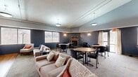 The Community Room at 9201 Nicollet Ave S in Bloomington, Minnesota, is a welcoming, single-floor multipurpose space designed to accommodate a variety of social gatherings and community activities. The main open area seamlessly blends a living room and dining room, featuring deep navy-blue walls accented by warm sconce lighting and pendant fixtures that create an inviting ambiance throughout. The living area is furnished with comfortable sofas and accent chairs arranged around area rugs atop wood-look flooring, providing a relaxed setting for conversation or casual gatherings. A cozy fireplace with a wood-tone mantle serves as a natural focal point, flanked by decorative accents. Several round dining tables with rolling chairs are distributed across the carpeted dining area, offering flexible seating arrangements for groups of varying sizes. Multiple windows throughout the space admit natural light and provide views of the surrounding outdoor landscape, while a glass door with sheer curtains offers direct outdoor access. Soaring ceilings in portions of the room enhance the sense of openness. The attached kitchen is outfitted with warm oak cabinetry, ample counter space, a full range, microwave, and sink — well-suited for catered events or community meals. Overall, the space balances functionality with a comfortable, communal atmosphere.