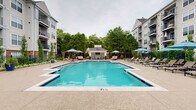 This outdoor amenity space centers around a generously sized swimming pool with vivid turquoise water and a classic tiled border, set within a well-maintained courtyard flanked by multi-story residential buildings. The pool deck is finished in textured concrete, providing ample space for lounging and circulation. Multiple chaise lounge chairs and shaded umbrella tables are arranged throughout the deck, offering both sun and shade options for residents.

At one end of the pool area sits a white-paneled cabana with a pitched roof and open-air design. Inside, the cabana features a coffered ceiling with a ceiling fan, wall-mounted flat-screen television, and comfortable outdoor seating including sofas and accent chairs arranged on a patterned area rug — creating a relaxed lounge atmosphere ideal for gatherings.

On the opposite end of the deck, a dedicated outdoor kitchen station is equipped with stainless steel grills and prep counters, providing a functional space for outdoor cooking and entertaining. Mature trees and manicured landscaping with ornamental plantings line the perimeter of the pool area, adding greenery and a sense of privacy. Decorative lamp posts contribute to the overall well-appointed character of the space.