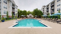 This outdoor amenity space centers around a generously sized swimming pool with vivid turquoise water and a classic tiled border, set within a well-maintained courtyard flanked by multi-story residential buildings. The pool deck is finished in textured concrete, providing ample space for lounging and circulation. Multiple chaise lounge chairs and shaded umbrella tables are arranged throughout the deck, offering both sun and shade options for residents.

At one end of the pool area sits a white-paneled cabana with a pitched roof and open-air design. Inside, the cabana features a coffered ceiling with a ceiling fan, wall-mounted flat-screen television, and comfortable outdoor seating including sofas and accent chairs arranged on a patterned area rug — creating a relaxed lounge atmosphere ideal for gatherings.

On the opposite end of the deck, a dedicated outdoor kitchen station is equipped with stainless steel grills and prep counters, providing a functional space for outdoor cooking and entertaining. Mature trees and manicured landscaping with ornamental plantings line the perimeter of the pool area, adding greenery and a sense of privacy. Decorative lamp posts contribute to the overall well-appointed character of the space.