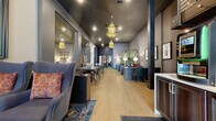 Located at 70 N High St in the heart of downtown Columbus, Ohio, this single-floor commercial space offers a generously proportioned, open layout that blends sophisticated design with an inviting atmosphere. The interior is defined by wide-plank light hardwood flooring that runs the length of the space, creating a warm contrast against the dramatic dark-painted ceilings in deep navy and charcoal tones. Ornate amber glass pendant chandeliers hang at intervals overhead, casting a warm, ambient glow throughout the room. The space is organized into distinct functional zones, including a long communal table with mixed seating—featuring cane-back and upholstered chairs with rich burgundy accents—alongside lounge areas with plush, patterned sofas and armchairs. A series of dark-framed workstation desks and credenzas line one side of the room, complemented by table lamps and lush greenery arrangements. Bold structural columns in matte black punctuate the space, adding architectural character. Large mirrored panels and framed artwork adorn the walls, enhancing the sense of depth and visual interest. A tiled entryway near the front adds a classic touch, while the open sightlines from front to back give the space an expansive, well-connected feel suited for a variety of uses.