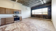 Located at 801 Second Ave N in Moorhead, MN, this one-bedroom, one-bathroom unit offers a distinctive loft-style aesthetic rooted in industrial character. The open living and kitchen area immediately draws the eye with its exposed concrete ceiling, visible ductwork and piping, and a striking original brick accent wall that speaks to the building's historic bones. Large, multi-pane windows flood the main living space with natural light, creating an airy atmosphere despite the unit's intimate footprint. The kitchen is equipped with stainless steel appliances — including a range, microwave, and refrigerator — alongside warm wood-toned cabinetry and a dishwasher, balancing functionality with style. A structural column anchors the open-plan space, adding to the loft's urban charm. The bedroom is a well-proportioned, quiet retreat with neutral walls and exposed ceiling details consistent with the rest of the unit. The bathroom features a walk-in shower stall, toilet, and vanity in a clean, understated design. A dedicated laundry area includes a stacked Whirlpool washer and dryer unit with adjacent wire shelving for added storage convenience. Throughout, the raw concrete-style flooring and industrial finishes give this unit a bold, creative character well-suited to those who appreciate urban, adaptive-reuse living.
