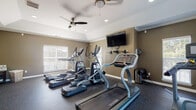 This fitness center offers a comprehensive workout environment designed to support a wide range of exercise routines. The space is spread across a single floor and is divided into distinct functional zones, making it easy to transition between cardio and strength training.

The cardio area is equipped with multiple treadmills, elliptical machines, and a StairMaster, all positioned near large windows fitted with blinds that allow for generous natural light. Ceiling fans overhead provide additional airflow and comfort during workouts. Wall-mounted flat-screen TVs are strategically placed for viewing during cardio sessions.

The strength training section features a power rack, a flat bench, and a well-organized dumbbell rack stocked with a full range of weights. A large wall mirror spans a significant portion of one wall, enhancing the sense of space while allowing users to monitor their form. Additional specialty cardio equipment, including an air bike, rounds out the offerings.

Throughout the facility, durable rubber flooring provides cushioning and traction. The walls are finished in a warm taupe tone with clean white crown molding and trim, lending a polished appearance. Recessed lighting complements the natural light from multiple windows and a glass-paneled exterior door, creating a bright and motivating atmosphere well-suited for daily fitness routines.