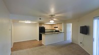 This single-floor residence offers a practical and comfortable open-concept layout that integrates the kitchen and living areas into one cohesive space. The kitchen is outfitted with light wood cabinetry, dark countertops, and black appliances, including a full-size refrigerator and range with overhead microwave. A peninsula counter extends into the living area, providing additional workspace and serving as a natural divider between the kitchen and the adjoining room. The living area features neutral carpet flooring, which contrasts with the hardwood-style flooring found in portions of the space. Ceiling fans are installed throughout, contributing to both comfort and air circulation. The walls are finished in a clean, neutral tone, and recessed lighting adds to the overall brightness of the interior. A hallway visible from the main living area leads to additional rooms, suggesting a well-organized floor plan. The space maintains a straightforward, functional design with consistent finishes throughout, making it both accessible and easy to navigate. Overall, the residence presents a clean, move-in ready environment suited for everyday living.