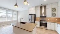 This thoughtfully designed single-floor space showcases a bright, open layout with a cohesive neutral color palette and light wood-look flooring throughout. The kitchen is a standout feature, centered around a large island with a warm quartz countertop and an integrated sink, illuminated by a pair of industrial-style pendant lights. White shaker cabinetry extends to the ceiling, offering generous storage, while a decorative geometric tile backsplash and a sleek stainless steel range hood add visual interest above the stainless steel gas range. A built-in microwave, stainless steel French door refrigerator, and dedicated display cabinetry with glass-front upper doors complete the well-equipped culinary space. Adjacent to the kitchen, a mudroom area features wall-mounted hooks, providing a practical drop zone at the entry. The living area is spacious and airy, anchored by a ceiling fan and separated from the private quarters by elegant sliding barn doors with frosted glass panels — a charming architectural detail that balances style and function. The bathroom offers a clean, spa-like atmosphere with a walk-in glass-enclosed shower featuring dual fixtures, a white vanity with warm countertop, and large-format tile flooring. Recessed lighting throughout ensures a bright, welcoming ambiance at every turn.
