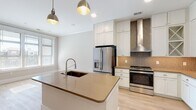 This thoughtfully designed single-floor space showcases a bright, open layout with a cohesive neutral color palette and light wood-look flooring throughout. The kitchen is a standout feature, centered around a large island with a warm quartz countertop and an integrated sink, illuminated by a pair of industrial-style pendant lights. White shaker cabinetry extends to the ceiling, offering generous storage, while a decorative geometric tile backsplash and a sleek stainless steel range hood add visual interest above the stainless steel gas range. A built-in microwave, stainless steel French door refrigerator, and dedicated display cabinetry with glass-front upper doors complete the well-equipped culinary space. Adjacent to the kitchen, a mudroom area features wall-mounted hooks, providing a practical drop zone at the entry. The living area is spacious and airy, anchored by a ceiling fan and separated from the private quarters by elegant sliding barn doors with frosted glass panels — a charming architectural detail that balances style and function. The bathroom offers a clean, spa-like atmosphere with a walk-in glass-enclosed shower featuring dual fixtures, a white vanity with warm countertop, and large-format tile flooring. Recessed lighting throughout ensures a bright, welcoming ambiance at every turn.