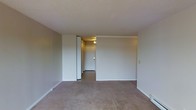 This single-floor space presents a straightforward and practical layout. The main living area is finished with wall-to-wall carpeting in a warm beige tone, providing a neutral foundation that suits a variety of furnishing styles. The walls are painted in a clean, light neutral shade, contributing to a bright and open feel throughout the room. Crown molding accents the upper portion of one wall, adding a subtle architectural detail to the otherwise understated interior. The ceiling is flat and finished in white, maintaining a clean and uniform appearance. A hallway visible from the main living area leads to additional rooms beyond, with overhead lighting illuminating the passage. White-trimmed doorframes and baseboards run throughout, offering a cohesive and tidy finish. The space is currently vacant, allowing for a clear view of the layout and finishes. Overall, the unit offers a clean, move-in-ready environment with a simple, functional design that provides a blank canvas for personalization.