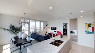 This single-floor residence offers a thoughtfully designed open-concept layout that seamlessly connects the kitchen, dining, and living areas. The kitchen is a standout feature, equipped with high-gloss white upper cabinetry, warm wood-toned lower cabinets, a stone mosaic tile backsplash, a gas range with a sleek hood, built-in oven, and a generously sized granite waterfall island with a double undermount sink. Large floor-to-ceiling windows flood the space with abundant natural light while framing sweeping city skyline views.

The living area is anchored by a bold black-and-white striped area rug and large windows that continue the panoramic urban vistas. Light hardwood-style flooring runs throughout, contributing to the space's bright, cohesive aesthetic.

Two bedrooms provide comfortable retreats, each featuring large windows with city views, soft neutral tones, and ample closet space. One bedroom is styled with a sleek black bed frame and monochromatic artwork, while the other features a plush upholstered headboard and botanical accents.

The bathrooms maintain a clean, contemporary finish with wood-grain vanities, white quartz countertops, large-format tile, and glass-enclosed showers. A bright, minimal hallway connects the private spaces, accented with a round mirror and sculptural wall-mounted planters.