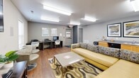 Located at 2205 S Washington St in Kokomo, Indiana, this single-floor community lounge offers a versatile and welcoming environment designed for both relaxation and productivity. The space features warm hardwood-style flooring throughout, neutral walls accented by a bold yellow feature wall, and bright fluorescent overhead lighting that keeps the area well-lit at all times.

The main living area is anchored by a yellow upholstered sofa with patterned accent pillows, complemented by swivel lounge chairs and a central coffee table set atop a decorative area rug. A wall-mounted flat-screen TV and a media console provide entertainment options, while high-top bistro tables with bar stools offer a casual seating alternative.

A dedicated workstation area along one wall features desks, computers, and a printer, making the space suitable for academic or professional use. A large chalkboard wall serves as a creative brainstorming or messaging surface, adding a collaborative element to the environment.

The kitchenette area includes cabinetry, a sink, and counter space, providing basic refreshment conveniences. Additional amenities include a popcorn machine, folding tables, and extra seating. Natural light filters in through multiple windows, and an emergency exit door ensures safety. The space is thoughtfully arranged to accommodate a variety of community activities.