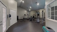 Located at 63 Turnberry Way in San Antonio, Texas, this single-story fitness center offers a functional and well-organized workout environment. The space features vaulted ceilings that lend an open, airy feel to the interior, complemented by recessed lighting and neutral gray walls throughout.

The workout area is thoughtfully divided into distinct exercise zones. A dedicated cardio section houses a treadmill, elliptical trainer, and stationary recumbent bike, all positioned in front of a full-length mirrored wall that visually expands the space. A cable weight machine provides options for strength training, while a flat weight bench and a punching bag round out the available equipment. Aerobic step platforms are also available for group-style exercises.

Durable rubber flooring covers the workout areas, providing cushioning and traction appropriate for exercise use. Large gridded windows allow natural light to filter into the space, while French doors open directly to an outdoor area with views of a pool, creating a seamless connection to the exterior. A carpeted transitional corridor near the entrance adds a subtle contrast in texture. Signage noting fitness center rules is posted near the entry, indicating this is a shared community amenity.
