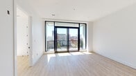 This thoughtfully designed single-floor urban residence showcases a clean, contemporary aesthetic throughout. The living area is defined by striking floor-to-ceiling black-framed sliding glass doors that open onto a private balcony, offering expansive views of the surrounding cityscape, including notable historic buildings on the horizon. An abundance of natural light floods the space through these large openings, complementing the light wood-toned luxury vinyl flooring found throughout the main living areas.

The kitchen features a modern, two-toned design with dark lower cabinetry contrasted against white square-tile backsplash that extends to the ceiling. Stainless steel appliances include a range with microwave above, a French door refrigerator, and a dishwasher. Open wood shelving adds a functional and stylish design element, while recessed and pendant lighting complete the space.

A separate bedroom area provides a calm retreat with clean white walls and generous natural light. The full bathroom is outfitted with large-format stone-look tile flooring, a full soaking tub with a white subway tile surround, a toilet, and matte black fixtures that add a refined, contemporary contrast to the bright white walls. The overall palette of whites, blacks, and natural wood tones creates a cohesive and modern living environment.