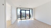 This thoughtfully designed single-floor urban residence showcases a clean, contemporary aesthetic throughout. The living area is defined by striking floor-to-ceiling black-framed sliding glass doors that open onto a private balcony, offering expansive views of the surrounding cityscape, including notable historic buildings on the horizon. An abundance of natural light floods the space through these large openings, complementing the light wood-toned luxury vinyl flooring found throughout the main living areas.

The kitchen features a modern, two-toned design with dark lower cabinetry contrasted against white square-tile backsplash that extends to the ceiling. Stainless steel appliances include a range with microwave above, a French door refrigerator, and a dishwasher. Open wood shelving adds a functional and stylish design element, while recessed and pendant lighting complete the space.

A separate bedroom area provides a calm retreat with clean white walls and generous natural light. The full bathroom is outfitted with large-format stone-look tile flooring, a full soaking tub with a white subway tile surround, a toilet, and matte black fixtures that add a refined, contemporary contrast to the bright white walls. The overall palette of whites, blacks, and natural wood tones creates a cohesive and modern living environment.