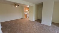 This single-floor space presents a clean, functional layout designed for comfortable everyday living. The main living area is generously sized, featuring wall-to-wall neutral carpeting that adds warmth underfoot. A ceiling fan with a light fixture is centrally mounted overhead, providing both air circulation and ambient lighting to the room. The walls are finished in a soft, light neutral tone, contributing to a bright and airy atmosphere. A closet with a standard door is conveniently located along one wall of the living area, offering practical storage solutions.

The living space opens directly into the kitchen through a wide, open passageway, creating a connected flow between the two areas. The kitchen features a tile or hard-surface floor, warm wood-toned cabinetry, and white appliances including a range and oven. An under-cabinet range hood is installed above the stove, and overhead lighting keeps the kitchen well-illuminated. The cabinetry provides ample storage and workspace for meal preparation. Overall, the space maintains a straightforward, practical aesthetic with neutral finishes that offer a versatile foundation for personalization.