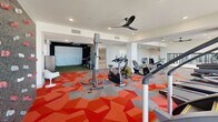 Located at 186 E Future Way in Draper, Utah, this single-floor fitness facility offers a generously sized, well-appointed space designed to accommodate a wide range of workout activities. The layout comprises two distinct exercise rooms and an additional flexible area, each thoughtfully designed to serve different training needs.

The main exercise areas are immediately striking, featuring bold geometric-patterned carpet in vibrant shades of red, orange, and gray that energizes the space. Large floor-to-ceiling windows line the perimeter, flooding the rooms with natural light and offering expansive outdoor views of the surrounding Draper landscape. The open floor plan is supported by structural columns and fitted with ceiling fans and recessed lighting throughout.

Strength training equipment, including Hammer Strength racks, cable machines, free weights, and benches, is distributed across the primary exercise zones, complemented by a dedicated cardio area housing treadmills, stationary bikes, ellipticals, and rowing machines. A climbing wall adds a unique recreational element to one section of the facility.

A separate studio room features smooth tile flooring, a wall-mounted television, mirrors, yoga mats, kettlebells, and a ballet barre, making it well-suited for group fitness classes or individual training sessions. The clean white walls and drop-tile ceiling create a calm, focused environment for varied workout disciplines.