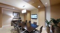 This conference room offers a well-appointed setting designed to support professional meetings and collaborative sessions. The space is anchored by a large, dark espresso-toned rectangular conference table surrounded by cushioned chairs with sleek chrome frames, providing comfortable seating for a sizable group. A wall-mounted flat-screen television serves as the focal point for presentations and video conferencing, positioned prominently for clear viewing from all seats. Overhead, a combination of recessed lighting and a central pendant fixture with a white drum shade creates a warm, well-balanced ambiance throughout the room. The warm beige and tan tones of the walls contribute to a professional yet inviting atmosphere. A framed art piece adorns one wall, adding a refined aesthetic touch, while a potted plant introduces a subtle natural element. Built-in cabinetry along one wall provides functional storage and workspace. The ceiling features a layered architectural detail with contrasting tones, adding visual depth and a polished, modern character to the room. Overall, the conference room is thoughtfully designed to balance functionality with a sophisticated, business-ready environment.