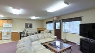 This single-floor space presents a welcoming and functional communal lounge area designed for comfort and relaxation. The room is anchored by a set of plush, cream-colored leather sofas and armchairs arranged around a glass-topped coffee table with an ornate metal frame, creating an inviting seating arrangement for gathering and conversation. A traditional fireplace with a wood mantel serves as a central focal point along one wall, adding warmth and a homey character to the space. Multiple windows fitted with white horizontal blinds and dark decorative valances allow generous natural light to fill the room, contributing to its bright and open atmosphere. The space is further illuminated by flush-mounted fluorescent ceiling fixtures set against a textured ceiling. Carpeted flooring in a deep, muted tone grounds the room and adds to its comfortable ambiance. A potted tropical plant near the entrance introduces a touch of greenery, while framed wall art adds a decorative accent. A door with sidelights provides access to the exterior, reinforcing the room's accessibility and connection to the outdoors. Overall, the space is practical, well-lit, and thoughtfully arranged to accommodate social interaction.