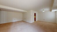This two-floor residence presents a straightforward and functional layout with neutral tones carried consistently throughout. The main living area is generously sized, featuring wall-to-wall beige carpeting, white walls, and a pendant light fixture overhead. A structural beam detail along the ceiling adds subtle architectural definition to the space. An opening leads directly into the kitchen, which is outfitted with warm oak cabinetry extending to the upper walls, white laminate countertops, and a full suite of appliances including a gas range, dishwasher, and refrigerator. The kitchen floor is finished in light-toned vinyl, contrasting with the carpeted areas of the home. The entry foyer welcomes natural light through glass panels flanking the front door, alongside a sliding glass door that provides access to the outdoors. Bedrooms continue the neutral, carpeted aesthetic, each offering closet access. The bathroom features white tile flooring, a single vanity with a large mirror, and clean, minimal fixtures. Overall, the residence maintains a cohesive, understated design that prioritizes practicality and livability across both floors.