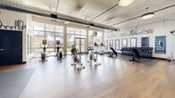 Located at 708 22nd St in Greeley, Colorado, this fitness center offers a generously sized, single-floor exercise space designed to support a wide range of workout routines. The room features warm hardwood flooring throughout, complemented by exposed ductwork and structural beams that lend an industrial character to the bright, open interior.

Large windows along one wall flood the space with natural light and provide views of the surrounding outdoor greenery, creating an inviting and energizing atmosphere. Pendant lighting fixtures are strategically placed to ensure consistent illumination across the workout area.

The fitness floor is comprehensively equipped with multiple cardio machines, including treadmills, upright bikes, and stationary cycling bikes. A dedicated free weights section features a full dumbbell rack and adjustable benches, while a functional training zone includes kettlebells, medicine balls, resistance equipment, and open floor space for dynamic exercises.

Framed wall mirrors are positioned throughout the space to assist with form and expand the sense of openness. A cityscape mural graphic adds a distinctive visual element to the walls, enhancing the modern aesthetic. Rubber mat flooring in designated strength and stretching zones provides safety and comfort. The well-organized layout ensures a smooth flow between workout zones, making this fitness center a practical and motivating environment for users of all fitness levels.
