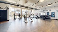 Located at 708 22nd St in Greeley, Colorado, this fitness center offers a generously sized, single-floor exercise space designed to support a wide range of workout routines. The room features warm hardwood flooring throughout, complemented by exposed ductwork and structural beams that lend an industrial character to the bright, open interior.

Large windows along one wall flood the space with natural light and provide views of the surrounding outdoor greenery, creating an inviting and energizing atmosphere. Pendant lighting fixtures are strategically placed to ensure consistent illumination across the workout area.

The fitness floor is comprehensively equipped with multiple cardio machines, including treadmills, upright bikes, and stationary cycling bikes. A dedicated free weights section features a full dumbbell rack and adjustable benches, while a functional training zone includes kettlebells, medicine balls, resistance equipment, and open floor space for dynamic exercises.

Framed wall mirrors are positioned throughout the space to assist with form and expand the sense of openness. A cityscape mural graphic adds a distinctive visual element to the walls, enhancing the modern aesthetic. Rubber mat flooring in designated strength and stretching zones provides safety and comfort. The well-organized layout ensures a smooth flow between workout zones, making this fitness center a practical and motivating environment for users of all fitness levels.