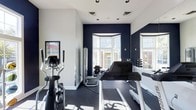 This single-floor fitness room is thoughtfully designed to support a comprehensive workout routine. The space features a crisp color palette of deep navy blue accent walls contrasted against clean white walls and ceiling, creating an energizing yet focused atmosphere. Natural light floods the room through large bay-style windows, offering views of the surrounding exterior and keeping the space bright and welcoming.

The flooring consists of dark rubber gym matting, practical for both equipment placement and exercise safety. Cardio equipment is well-represented, with multiple treadmills and an elliptical machine positioned to take advantage of the natural light. A cable strength machine is also present, complementing the cardiovascular offerings. A dumbbell rack with multiple weight increments lines one wall, catering to free-weight training needs.

Recessed and track lighting supplements natural light for consistent illumination throughout the day. A workout reference chart is mounted on the wall, providing exercise guidance. A large mirror expands the sense of space and allows users to monitor their form. The overall ambiance is clean, functional, and modern.