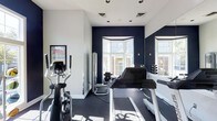 This single-floor fitness room is thoughtfully designed to support a comprehensive workout routine. The space features a crisp color palette of deep navy blue accent walls contrasted against clean white walls and ceiling, creating an energizing yet focused atmosphere. Natural light floods the room through large bay-style windows, offering views of the surrounding exterior and keeping the space bright and welcoming.

The flooring consists of dark rubber gym matting, practical for both equipment placement and exercise safety. Cardio equipment is well-represented, with multiple treadmills and an elliptical machine positioned to take advantage of the natural light. A cable strength machine is also present, complementing the cardiovascular offerings. A dumbbell rack with multiple weight increments lines one wall, catering to free-weight training needs.

Recessed and track lighting supplements natural light for consistent illumination throughout the day. A workout reference chart is mounted on the wall, providing exercise guidance. A large mirror expands the sense of space and allows users to monitor their form. The overall ambiance is clean, functional, and modern.