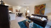This single-story residence showcases an open-concept layout that seamlessly connects the living, dining, and kitchen areas into one cohesive living space. The living room is anchored by a dark upholstered sofa adorned with decorative pillows, positioned atop a vibrant teal area rug that adds a bold pop of color against the neutral carpeted flooring. A decorative trunk serves as a coffee table, complementing the eclectic artwork displayed on the walls. A tall arc floor lamp provides warm ambient lighting to the seating area.

The dining area sits adjacent to the living space and accommodates a round table with matching dark chairs, positioned beneath a ceiling fan with integrated lighting. Neutral-toned walls and light carpet throughout create a cohesive, airy feel across the open floor plan.

A kitchen counter with a raised breakfast bar is visible at the edge of the space, offering a casual dining or workspace option. A hallway visible in the background leads to additional rooms, with a glimpse of what appears to be a bathroom vanity with warm lighting, suggesting well-distributed living spaces throughout the home.