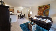 This single-story residence showcases an open-concept layout that seamlessly connects the living, dining, and kitchen areas into one cohesive living space. The living room is anchored by a dark upholstered sofa adorned with decorative pillows, positioned atop a vibrant teal area rug that adds a bold pop of color against the neutral carpeted flooring. A decorative trunk serves as a coffee table, complementing the eclectic artwork displayed on the walls. A tall arc floor lamp provides warm ambient lighting to the seating area.

The dining area sits adjacent to the living space and accommodates a round table with matching dark chairs, positioned beneath a ceiling fan with integrated lighting. Neutral-toned walls and light carpet throughout create a cohesive, airy feel across the open floor plan.

A kitchen counter with a raised breakfast bar is visible at the edge of the space, offering a casual dining or workspace option. A hallway visible in the background leads to additional rooms, with a glimpse of what appears to be a bathroom vanity with warm lighting, suggesting well-distributed living spaces throughout the home.
