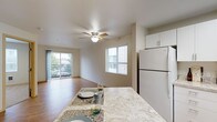 This single-floor residence is designed with an open, functional layout that connects the kitchen and living areas seamlessly. The kitchen is well-appointed with white cabinetry, light granite-style countertops, white appliances including a refrigerator, electric range with overhead microwave, and dishwasher. A central island provides additional counter space and doubles as a casual dining surface. Wood-style flooring runs throughout the kitchen and living areas, contributing to a clean, cohesive aesthetic. A skylight-style fixture and window above the sink enhance natural light in the kitchen.

The open living area features a ceiling fan with light kit and benefits from multiple windows fitted with white blinds. A sliding glass door leads to a private balcony with outdoor views. Adjacent to the living space, a carpeted bedroom offers a quiet retreat, complete with a window and wall-mounted heating unit. A full bathroom includes a white vanity with a marble-style countertop, a framed mirror, and hardwood-style flooring.

A dedicated laundry closet houses a stacked washer and dryer unit, adding everyday convenience. Throughout the space, neutral walls, light flooring, and consistent hardware create a cohesive, move-in-ready environment.