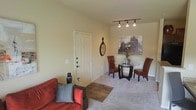 This single-floor space combines a living area and dining nook within an open, efficiently designed layout. The living area features plush carpeting and a warm color palette, with soft yellow-toned walls creating an inviting atmosphere. A bold red sofa anchors the seating area, complemented by a decorative area rug that adds texture and visual contrast to the floor.

The dining area sits adjacent to the living space, furnished with a round glass-top table and rich red leather chairs, offering a compact yet functional setting for meals. Overhead, a track lighting fixture provides direct, adjustable illumination, enhancing the usability of the space. A large framed cityscape artwork serves as a focal point on the wall, adding a decorative element that draws the eye.

A solid white paneled entry door with a deadbolt provides secure access to the unit. Decorative wall accents, including a round clock and small accent pieces, add personality throughout the space. The overall ambiance is warm and intimate, making efficient use of the available footprint while maintaining a comfortable and functional living environment.