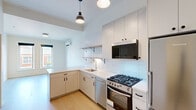 This single-floor unit showcases a thoughtfully designed, modern interior with a clean, minimalist aesthetic. The open-concept layout seamlessly connects the living area to the kitchen, creating a sense of spaciousness and flow throughout. Light hardwood flooring runs continuously across the unit, adding warmth and cohesion to the bright, neutral color palette.

The kitchen is a standout feature, outfitted with flat-front cabinetry in a soft gray-white finish, white subway tile backsplash extending to the ceiling, and open black metal shelving for both function and style. Stainless steel appliances include a gas range, built-in microwave, dishwasher, and a full-size Blomberg refrigerator. White quartz countertops and a stainless undermount sink complete the polished look. Industrial-style pendant lights overhead add a contemporary touch.

The living area is open and airy, benefiting from natural light that filters through large windows offering views of the surrounding brick-facade neighborhood buildings. A wall-mounted split-unit HVAC system provides efficient climate control throughout the space.

The bathroom, visible in the 3D model, features a clean white vanity and toilet. The overall ambiance of the unit is bright, modern, and move-in ready, with every detail reflecting contemporary urban design.