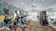 The Crowne at James Landing Fitness Center is a well-appointed, single-story facility designed to support a comprehensive workout experience. The main cardio area features soaring vaulted ceilings with recessed lighting and ceiling fans, creating an open, airy atmosphere. A row of treadmills, ellipticals, and stair climbers lines the space, oriented toward large windows that invite natural light and outdoor views. A bold teal, yellow, and white striped accent wall adds a vibrant, modern aesthetic, complemented by neutral herringbone-patterned carpet tiles throughout.

A dedicated strength training zone houses a multi-station cable machine, a full dumbbell rack, and additional resistance equipment, providing ample options for resistance-based workouts. Adjacent to the main floor, a functional training studio offers floor-to-ceiling mirrors, TRX suspension trainers, heavy punching bags mounted to a wall-mounted rack, and medicine balls, making it ideal for dynamic and circuit-style training. Natural light from multiple windows enhances this space.

A separate cardio nook features a spin bike, elliptical, and wall-mounted flat-screen TV, offering a more intimate workout setting. Together, these zones create a versatile, well-lit fitness environment suited to a wide range of exercise preferences.