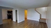 This three-floor space is designed with functionality and practicality in mind. The main level, visible in the image, features warm hardwood-style flooring that runs throughout, complementing the neutral grey walls and crisp white trim. A textured popcorn ceiling adds a classic touch overhead.

The open layout of the main level provides a generous living area, with a built-in closet offering convenient storage. A doorway leads to an adjacent room, which features warm-toned walls and continues the hardwood-style flooring, suggesting a seamlessly connected living space. A staircase positioned along the far wall provides access to the upper and lower floors, with clean lines and a carpeted tread design.

Baseboard heating units are visible along the lower walls, ensuring efficient climate control throughout the space. Recessed electrical outlets and cable hookups are thoughtfully placed throughout the main level. The overall ambiance is clean, neutral, and move-in ready, offering a blank canvas that accommodates a variety of interior design preferences across all three levels.