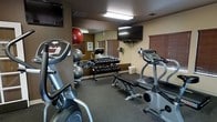 This single-level fitness room offers a practical and functional space for exercise, thoughtfully arranged to maximize its compact footprint. The room features neutral-toned walls with dark brown accents, complemented by overhead fluorescent lighting that ensures a well-lit environment. A large wall mirror along one side visually expands the space while allowing users to monitor their form during workouts.

The cardio section includes an upright stationary bike and an elliptical trainer, both equipped with digital consoles for tracking workout metrics. A wall-mounted flat-screen television is positioned for easy viewing from the cardio equipment, enhancing the exercise experience. For strength training, a multi-tiered dumbbell rack holds a comprehensive range of free weights, accompanied by a flat workout bench for versatile exercise options. Exercise balls, including a red stability ball and a grey medicine ball, add further variety to available workouts.

The flooring consists of durable, dark rubberized material suited for high-traffic fitness use. Slatted window blinds provide adjustable natural light and privacy. The overall ambiance is straightforward and utilitarian, prioritizing function and accessibility.