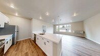 Located at 1779 Village Ln SE in Port Orchard, Washington, this single-floor 2-bedroom, 2-bathroom home offers a clean, modern design with consistent wood-look flooring and neutral warm-toned walls throughout. The open kitchen is a standout feature, equipped with stainless steel appliances including a range, microwave, and refrigerator, complemented by crisp white shaker cabinetry with black hardware and a generous quartz countertop with subtle gold veining that doubles as a breakfast bar. The living area is bright and welcoming, with a large picture window offering views of mature trees and the surrounding greenery. The primary bedroom is a comfortable, well-proportioned retreat with multiple windows that bring in natural light and views of the outdoors. The second bedroom also features tall windows and shares the same warm, neutral palette. Both bathrooms are finished with white vanities with black hardware; one includes a tub-shower combination, while the other incorporates a stacked washer and dryer unit for added convenience. White panel interior doors and clean baseboards lend a cohesive, polished look throughout the home. The efficient, well-organized layout maximizes every square foot, making this residence a practical and attractive option in the Port Orchard area.