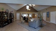This single-floor space makes a striking first impression with its dramatically vaulted ceilings that rise to a sharp peak, creating an expansive sense of volume throughout the main living area. Two ornate chandeliers hang in tandem, casting warm light across the open-concept floor plan and adding a refined, decorative touch to the space. A clerestory window positioned high on the accent wall draws in additional natural light, further enhancing the airy atmosphere.

The main living area features light-toned hardwood flooring that extends seamlessly throughout, anchored by a plush area rug beneath a generously sized sectional sofa. Warm earth-toned walls provide a welcoming backdrop that complements the neutral furnishings. A dedicated dining area sits nearby, furnished with a dark wood table and upholstered chairs, creating a natural separation of function within the open layout.

Large interior windows and glass-paned doors connect the main area to adjacent rooms, maintaining a sense of openness and visual continuity throughout the space. Additional rooms are visible beyond, suggesting further functional areas. A bookshelf and decorative accents, including tall floral arrangements, contribute to the overall polished and inviting ambiance of this well-appointed space.