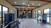 This modern fitness center offers a thoughtfully designed single-floor layout that balances functionality with aesthetic appeal. The space is anchored by light hardwood-style flooring that lends a clean, contemporary feel throughout. Decorative wood beam accents along the ceiling add a warm architectural contrast to the otherwise crisp, neutral interior. Recessed lighting, complemented by ceiling fans, ensures the space remains well-lit and comfortable during workouts. Large windows on multiple walls flood the gym with natural light while offering views of the surrounding exterior, creating an open and inviting atmosphere. The fitness floor is comprehensively outfitted with a full dumbbell rack, cable machines, resistance training equipment, and a variety of cardio machines, making it suitable for a broad range of fitness routines. Full-length mirrors are strategically placed to support form monitoring and to visually expand the space. The layout is thoughtfully organized to allow ease of movement between equipment zones. Overall, the fitness center presents a polished and professional environment ideal for daily exercise, combining practical amenities with an upscale design sensibility.