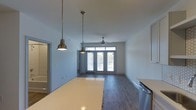 Located at 1817 Sir Tyler Dr in Wilmington, NC, this single-story space showcases a thoughtfully designed open-concept layout that seamlessly connects the kitchen and living areas. The kitchen features light-toned countertops and is equipped with stainless steel appliances, while a pair of brass pendant lights hang overhead, adding a warm, industrial-modern touch to the space.

The living area extends naturally from the kitchen, highlighted by rich, dark hardwood-style flooring that runs throughout. A ceiling fan centered in the living space provides both function and a subtle design element. Large French doors at the far end of the room flood the interior with abundant natural light and provide direct access to an outdoor area, creating a bright and airy atmosphere.

The clean white walls and neutral color palette throughout contribute to a fresh, modern aesthetic. The open sightlines from the kitchen counter through to the French doors enhance the sense of spaciousness. Additional doorways visible along the side of the space suggest further rooms, completing this well-appointed, single-level layout.