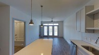Located at 1817 Sir Tyler Dr in Wilmington, NC, this single-story space showcases a thoughtfully designed open-concept layout that seamlessly connects the kitchen and living areas. The kitchen features light-toned countertops and is equipped with stainless steel appliances, while a pair of brass pendant lights hang overhead, adding a warm, industrial-modern touch to the space.

The living area extends naturally from the kitchen, highlighted by rich, dark hardwood-style flooring that runs throughout. A ceiling fan centered in the living space provides both function and a subtle design element. Large French doors at the far end of the room flood the interior with abundant natural light and provide direct access to an outdoor area, creating a bright and airy atmosphere.

The clean white walls and neutral color palette throughout contribute to a fresh, modern aesthetic. The open sightlines from the kitchen counter through to the French doors enhance the sense of spaciousness. Additional doorways visible along the side of the space suggest further rooms, completing this well-appointed, single-level layout.