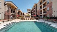 Located at 600 St Mary's St in Raleigh, NC, this thoughtfully designed outdoor amenity courtyard is set within a multi-story mixed-use development featuring a striking blend of brick, stucco, and stone architecture. The space is organized around a central resort-style swimming pool, flanked by rows of lounge chairs and shaded by contemporary sail canopies, creating an inviting atmosphere for relaxation. A prominent stone-clad water feature serves as a focal point, with a glass-enclosed fire feature nearby that provides warmth and ambiance for evening gatherings. Throughout the courtyard, raised brick planter beds overflow with lush, colorful landscaping — including tropical palms, ornamental shrubs, flowering plants, and manicured greenery — giving the space a vibrant, garden-like character. Multiple seating areas are distributed across the wide-format tile patio, furnished with wicker sofas, cushioned chairs, and dining sets shaded by bold red umbrellas, accommodating both social gatherings and quiet relaxation. Surrounding balconies on the upper floors of the building overlook the courtyard, reinforcing the sense of an enclosed, community-centered outdoor retreat. The open-sky setting ensures abundant natural light throughout the day.