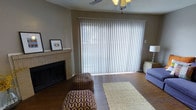 This single-story space in Arlington, Texas offers a comfortable and functional layout centered around a welcoming living area. The living room is highlighted by a classic brick fireplace, serving as a focal point along one wall and adding warmth and character to the space. Dark hardwood flooring runs throughout, providing a rich, grounded aesthetic that contrasts nicely with the neutral taupe wall tones and white trim.

A large sliding glass door spans nearly the full width of one wall, fitted with vertical blinds that allow for adjustable natural light and privacy control. This expansive glass feature fills the room with daylight and provides a visual connection to the outdoors. A ceiling fan with a light fixture is centrally positioned, ensuring comfortable air circulation throughout the room.

The textured ceiling adds a traditional touch consistent with the overall design sensibility of the space. The living area comfortably accommodates seating and accent pieces, as evidenced by the patterned ottomans and area rug that define the gathering space. Overall, the room presents a relaxed, livable atmosphere well-suited for everyday comfort in a North Texas setting.