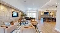 Located at 6000 Eldorado Pkwy in Frisco, TX, this single-story community amenity space offers a generous, well-appointed layout designed for both socializing and productivity. The main lounge area anchors the space with a striking textured accent wall, a wall-mounted TV, and comfortable seating arrangements set atop a bold geometric area rug — all bathed in natural light streaming through large windows with sheer curtains. Adjacent to the lounge is a sophisticated wet bar featuring an expansive marble-topped island with bar seating, navy lower cabinetry, a subway tile backsplash, open display shelving, and dramatic mixed-metal pendant lighting overhead. A dedicated game room provides a lively counterpoint, complete with a ping pong table, artwork, and direct access to an outdoor patio. A coworking nook offers built-in wood shelving with integrated lighting, white stone desks, and leather task chairs — ideal for focused work. Private study rooms with built-in desks, glass-panel doors, and acoustic flooring provide quiet individual workspaces. Light hardwood flooring flows consistently throughout, tying together the various zones into a cohesive, modern, and inviting environment.